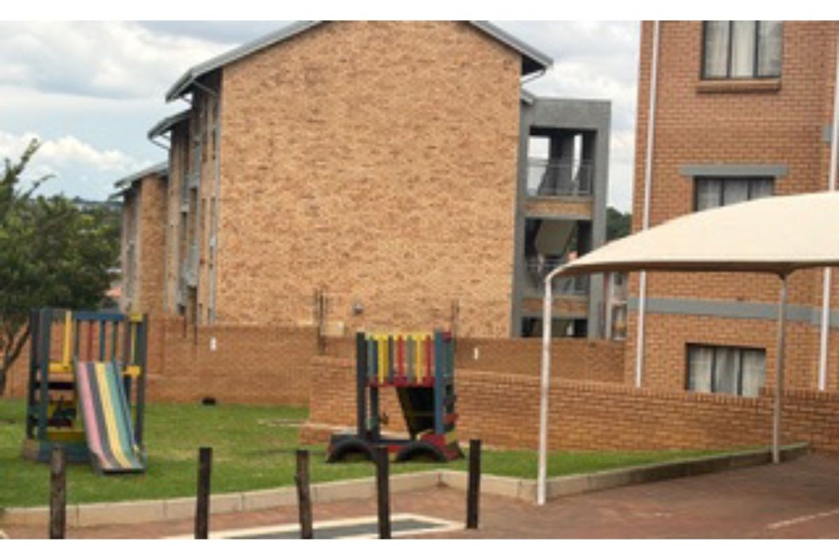 Exterior view of an apartment complex featuring a communal garden area with a playground and a shaded parking or seating structure.