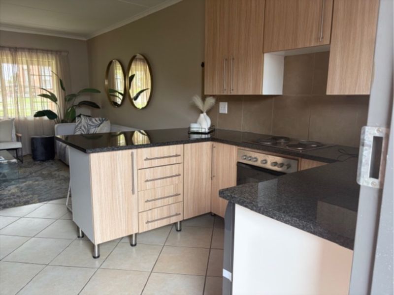 Open-plan kitchen featuring light wood cabinetry, granite countertops, and integrated electric cooking appliances, flowing into a comfortable living area with tiled flooring.