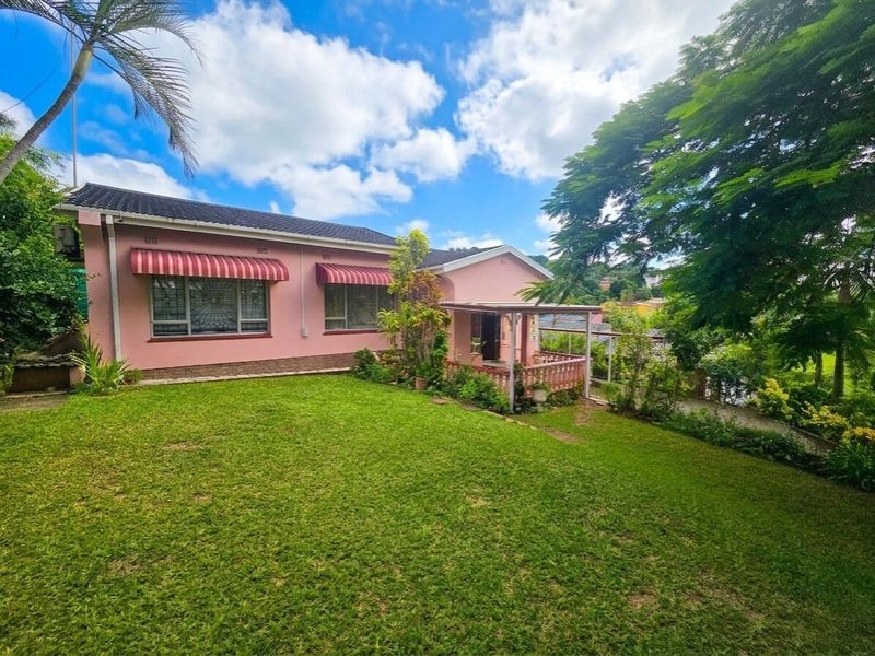 A view of the rear elevation of a pink house featuring a covered patio, security-barred windows with awnings, and a well-maintained lawn.