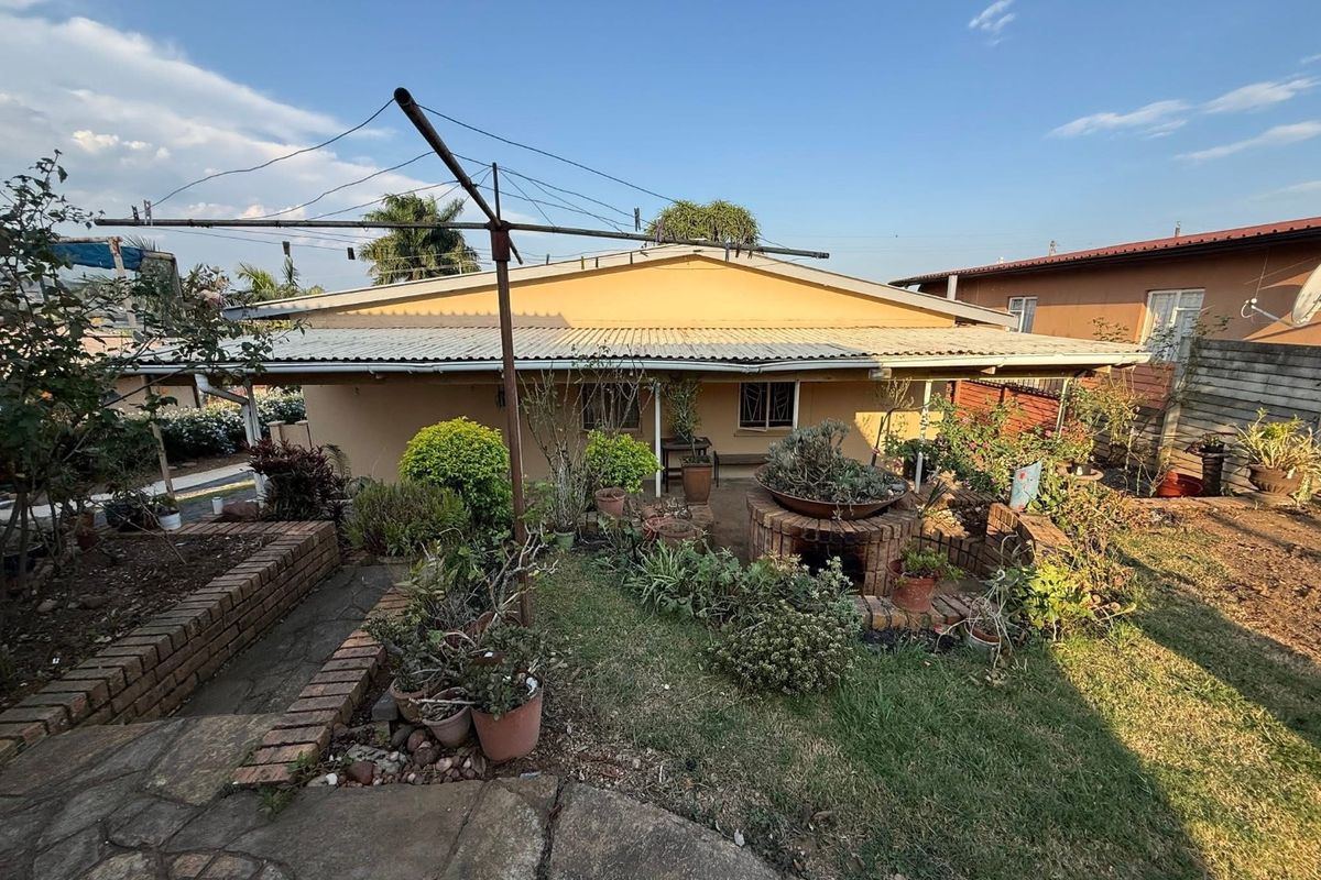 Rear view of the property featuring a corrugated roof, brick retaining walls, a stone pathway, and a built-in brick braai area in the garden.