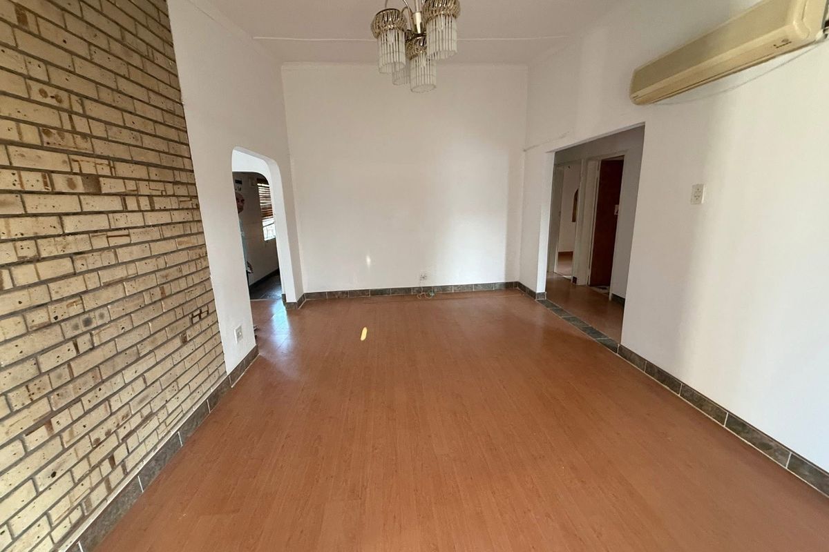 An empty living room featuring laminate flooring, an exposed brick feature wall, and a wall-mounted air conditioning unit.