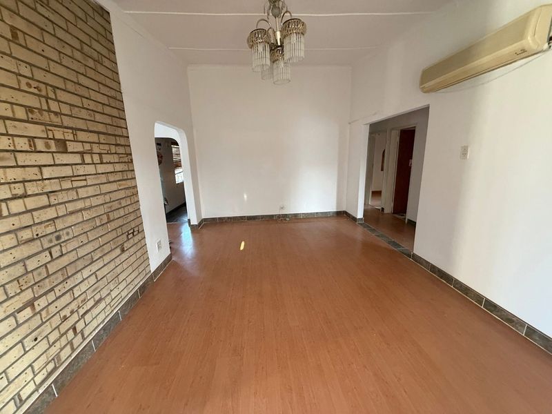 An empty living room featuring laminate flooring, an exposed brick feature wall, and a wall-mounted air conditioning unit.