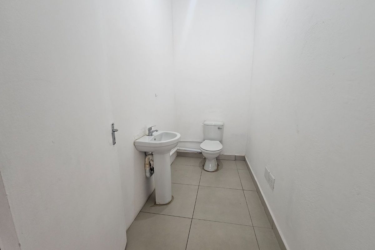 A functional, minimalist bathroom featuring a pedestal washbasin and toilet with neutral floor tiling and plain white walls.