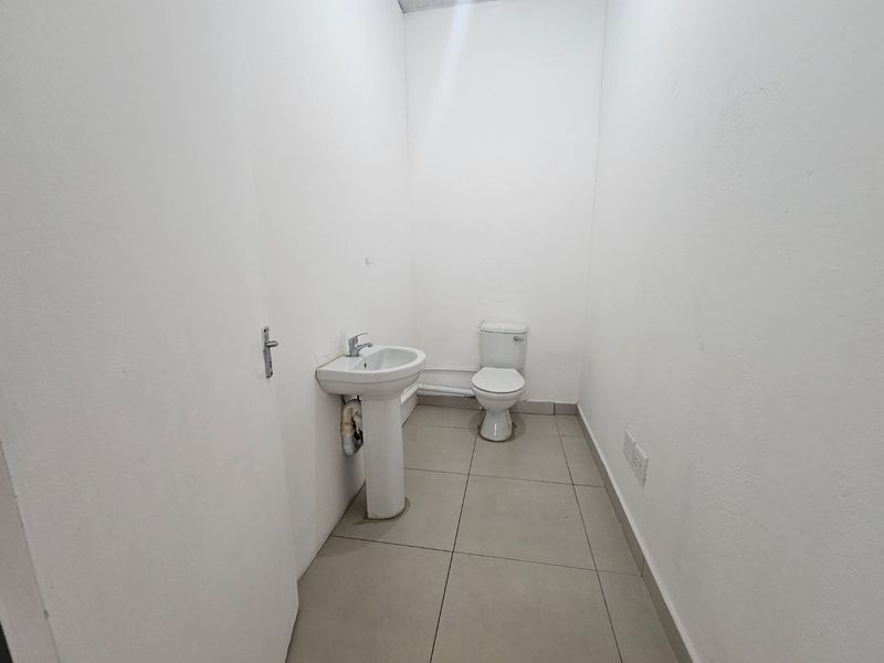 A functional, minimalist bathroom featuring a pedestal washbasin and toilet with neutral floor tiling and plain white walls.