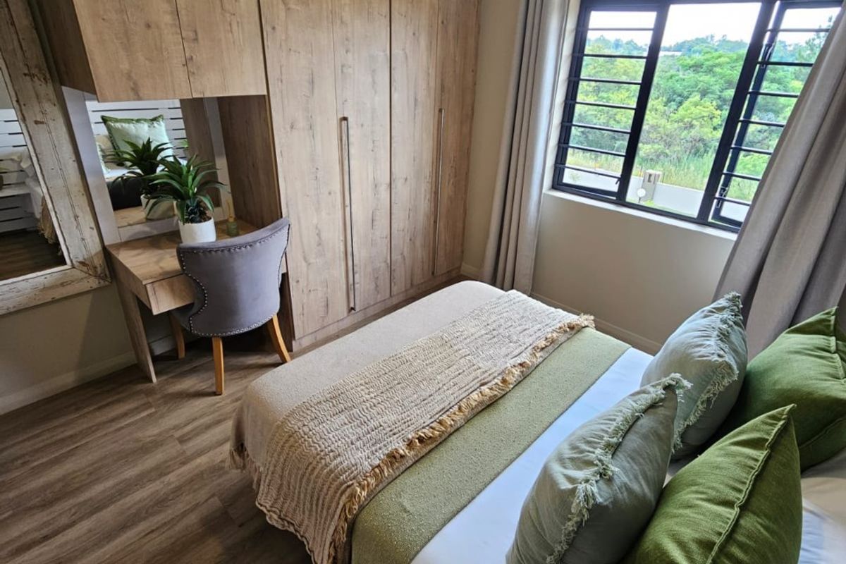 A well-maintained bedroom featuring built-in wooden cupboards with an integrated vanity area, laminate flooring, and a large window overlooking greenery.
