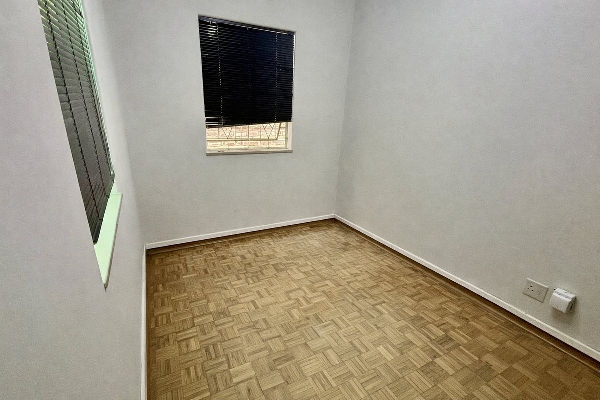 A clean, empty bedroom featuring classic parquet flooring, neutral walls, and windows fitted with venetian blinds and security bars.