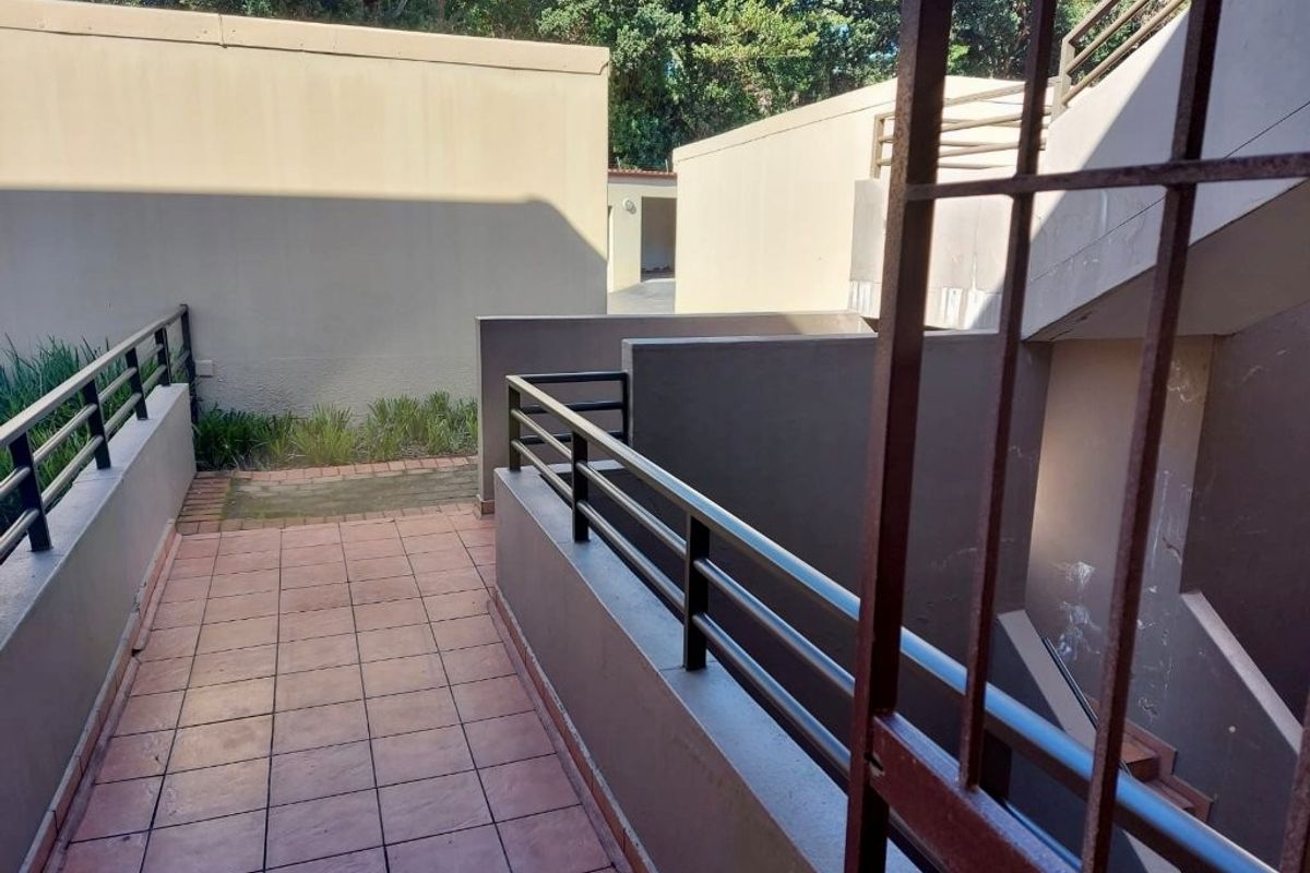 An exterior rear view of an apartment complex featuring a tiled patio area, metal balustrades, and an external staircase.