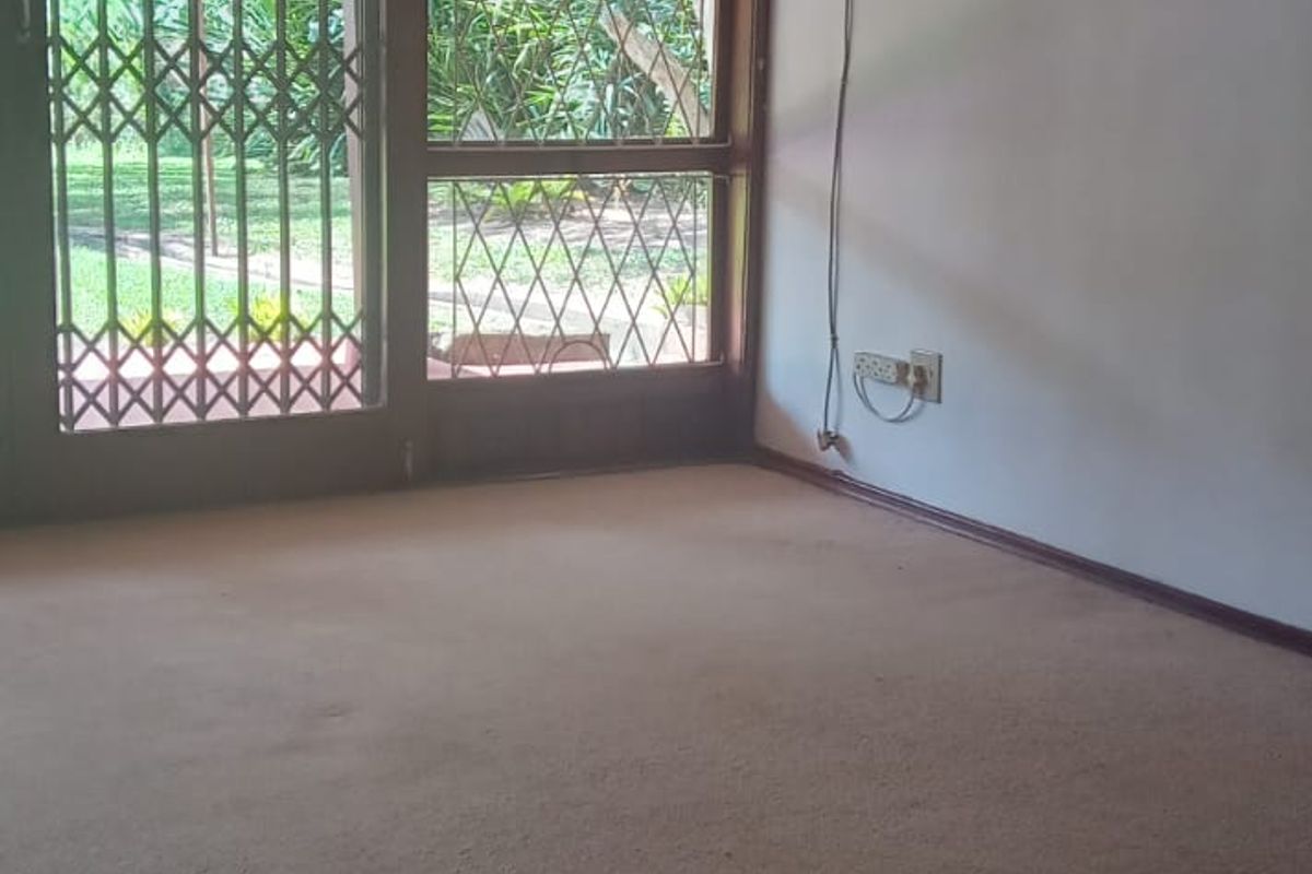 An empty living room featuring carpet flooring, a ceiling fan, and a window-mounted air conditioning unit with access to the garden through a sliding door.
