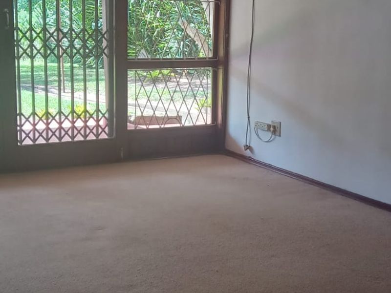 An empty living room featuring carpet flooring, a ceiling fan, and a window-mounted air conditioning unit with access to the garden through a sliding door.