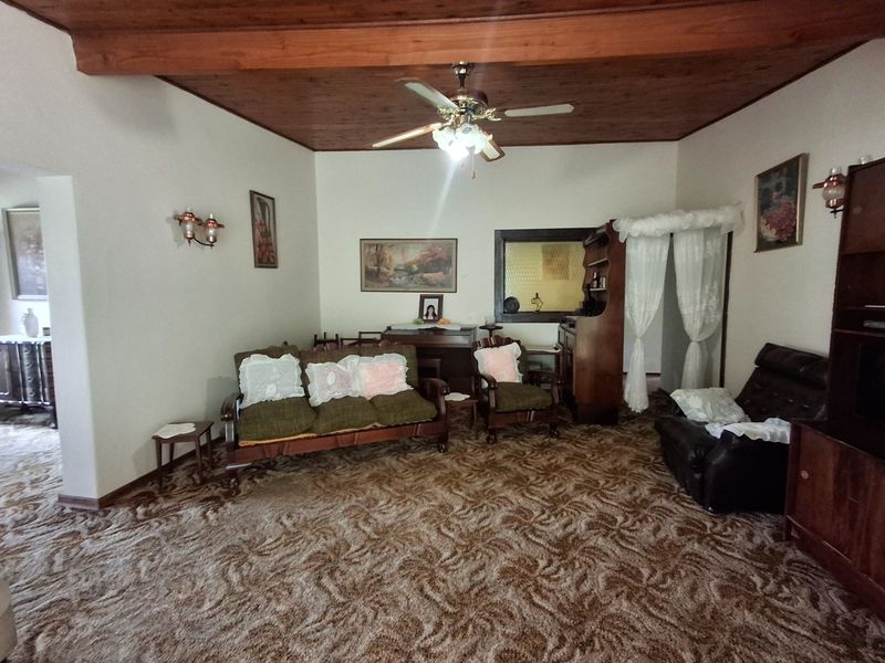 A living room featuring an exposed timber ceiling, ceiling fan, and wall sconces, with dated carpet flooring throughout.