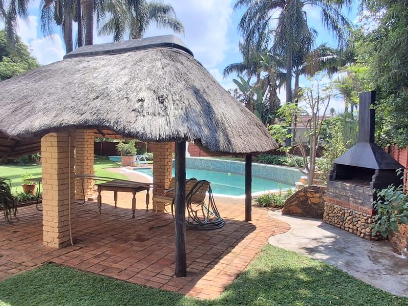 Outdoor entertainment area featuring a thatched lapa, brick paving, a built-in braai, and a swimming pool set within a landscaped garden.