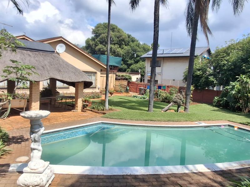 Outdoor entertainment area featuring a swimming pool, a traditional thatched lapa, and a landscaped garden with mature palm trees.