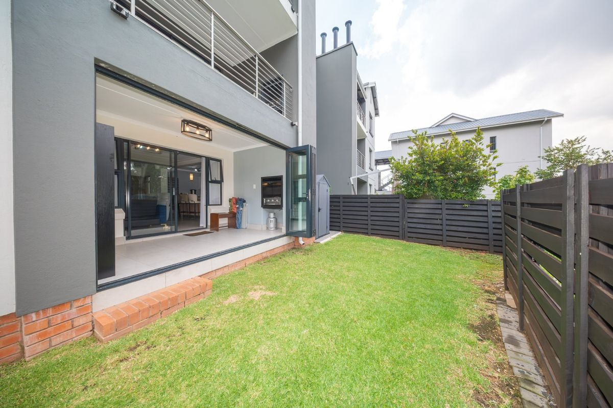 A modern townhouse rear exterior featuring a covered patio with a built-in braai, bi-fold doors, and a private lawn area with timber fencing.