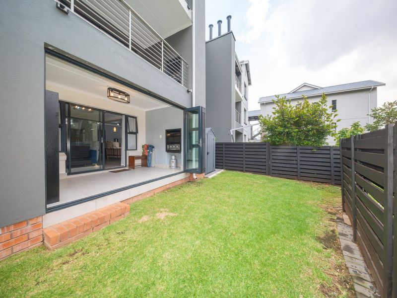 A modern townhouse rear exterior featuring a covered patio with a built-in braai, bi-fold doors, and a private lawn area with timber fencing.