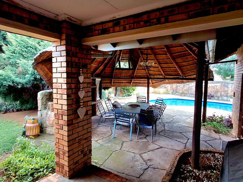 Covered outdoor patio featuring a thatched roof, brick pillars, and stone paving, overlooking a swimming pool and garden area.