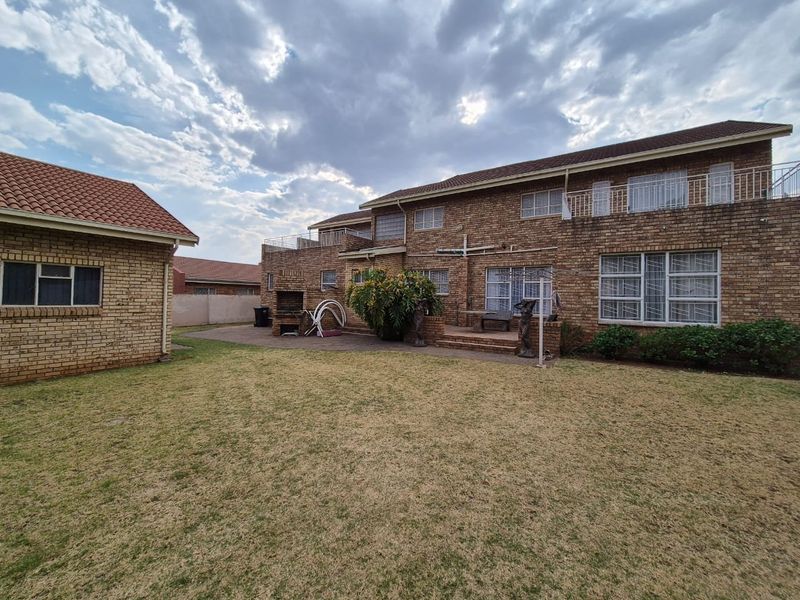 Rear view of a double-story brick house featuring a paved patio, built-in braai, upper-level balcony, and a spacious garden area.