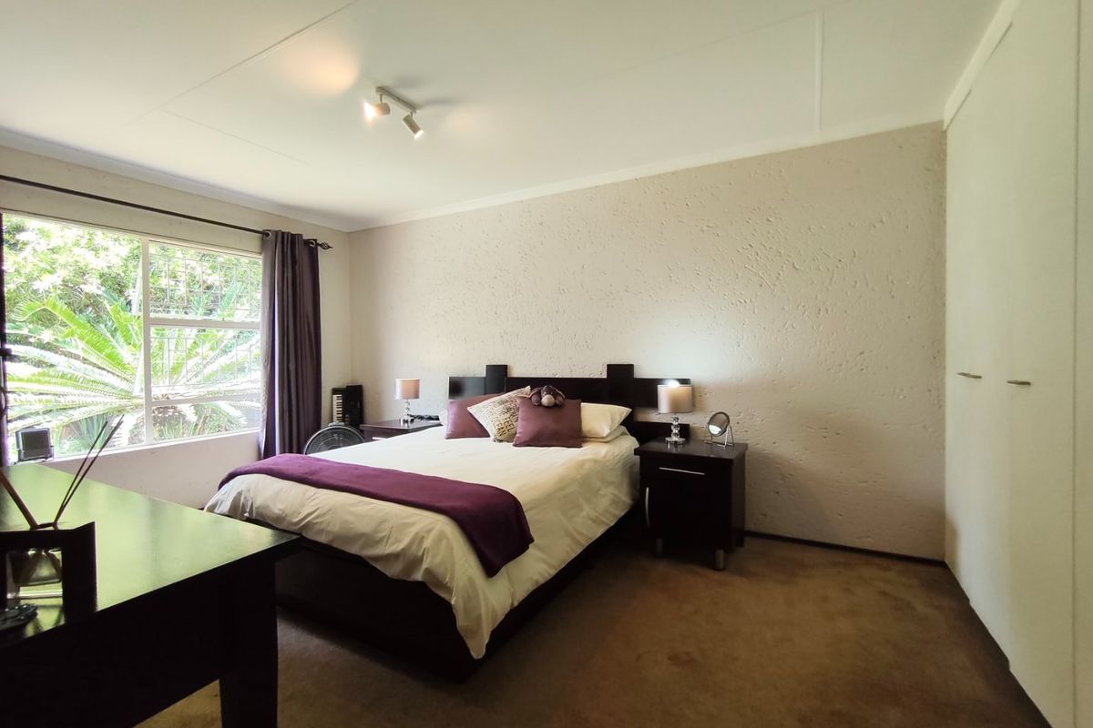 A well-proportioned bedroom featuring built-in cupboards, neutral textured walls, carpeted flooring, and a large window with security bars overlooking the garden.