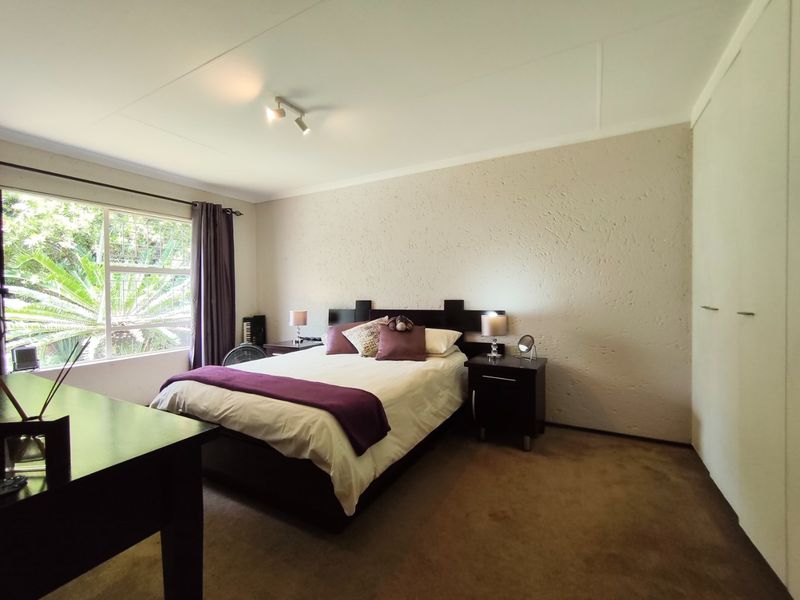 A well-proportioned bedroom featuring built-in cupboards, neutral textured walls, carpeted flooring, and a large window with security bars overlooking the garden.