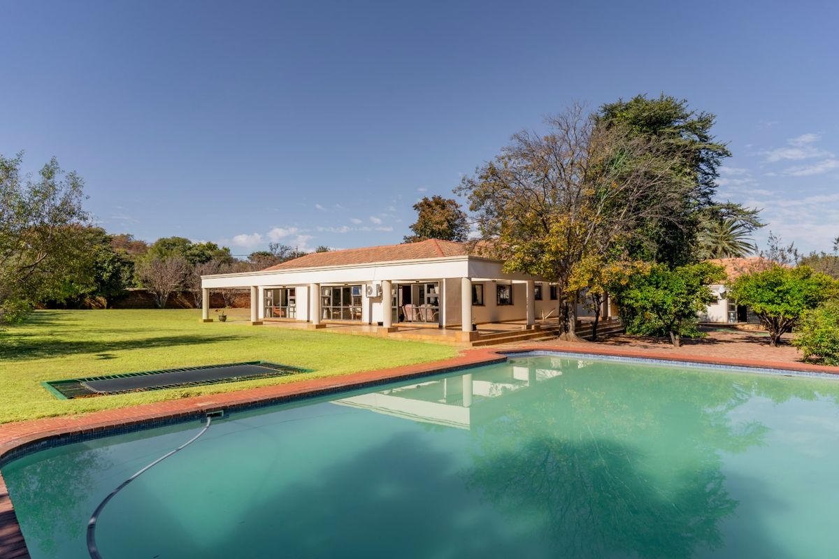 A spacious rear exterior view featuring a large swimming pool, a covered patio area, and a well-maintained garden with mature trees.