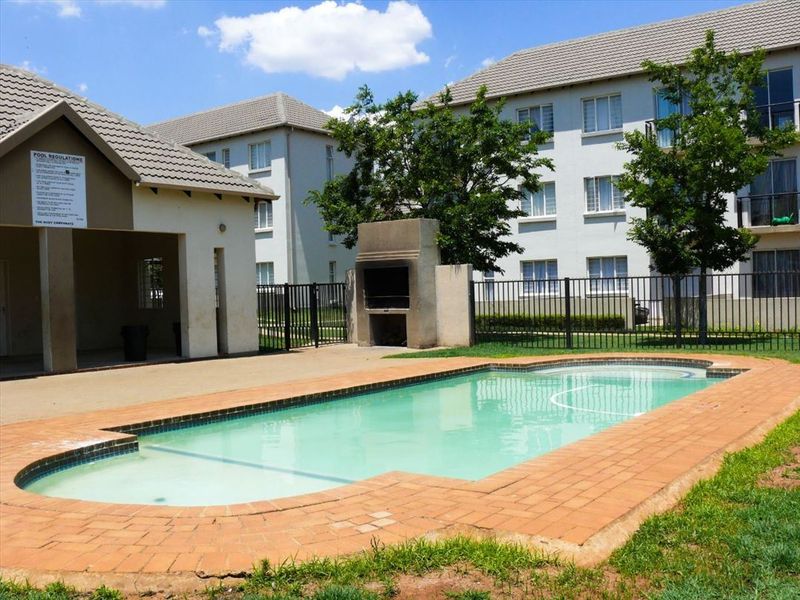 Communal swimming pool area featuring a built-in braai, covered patio space, and secure metal perimeter fencing within an apartment complex.