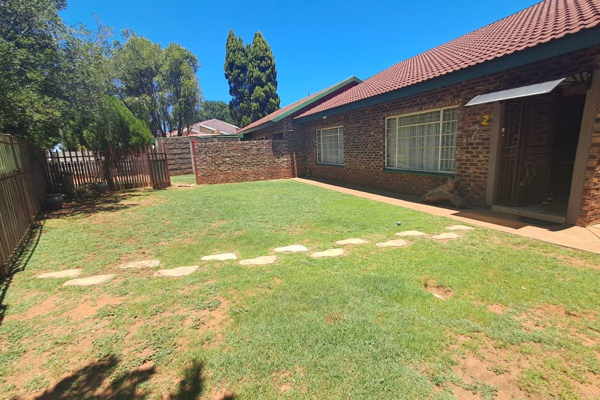 A rear exterior view of a brick house featuring a lawn, stone pathway, and secure boundary fencing under a clear blue sky.
