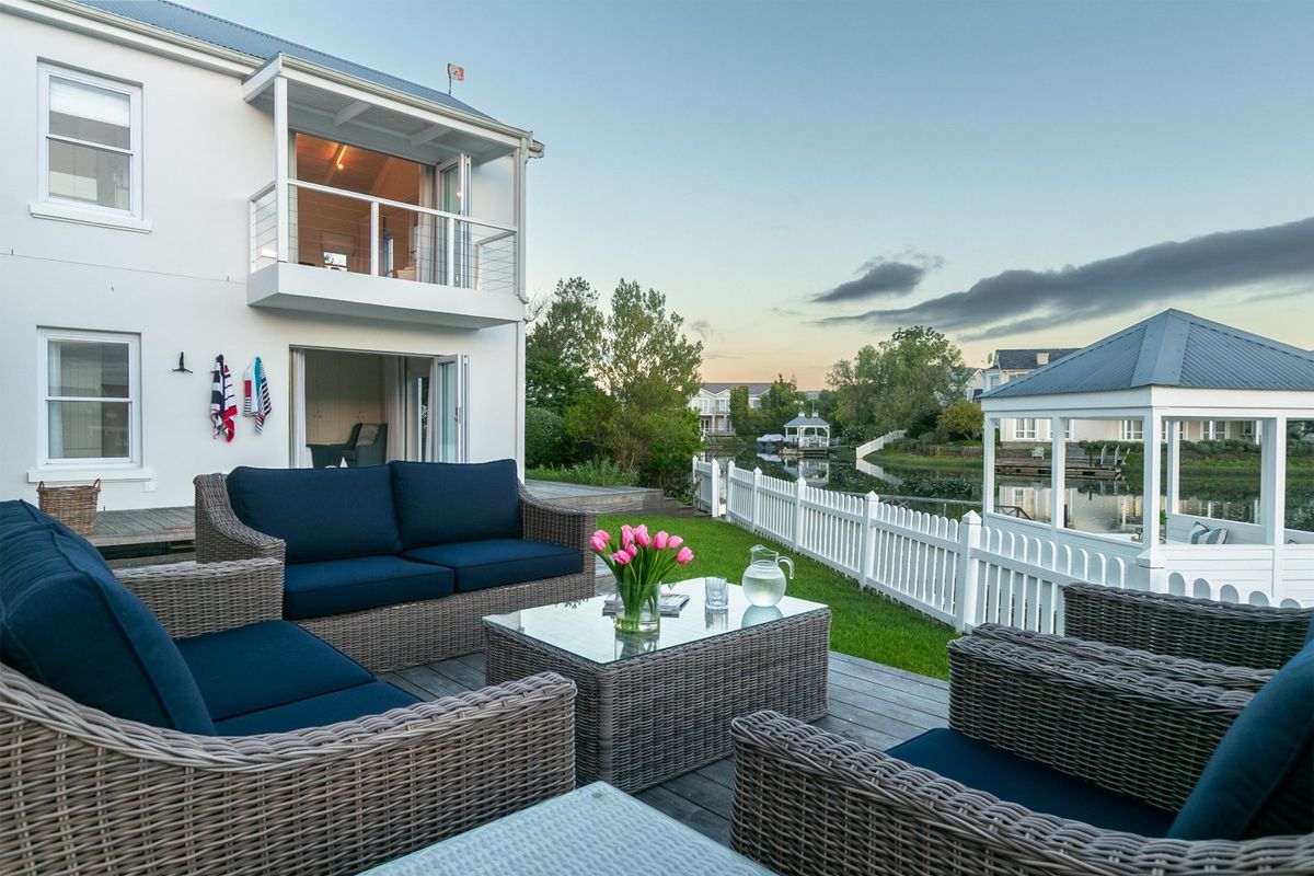 A well-maintained rear patio area featuring timber decking, a white picket fence, and a balcony overlooking a tranquil canal and gazebo.