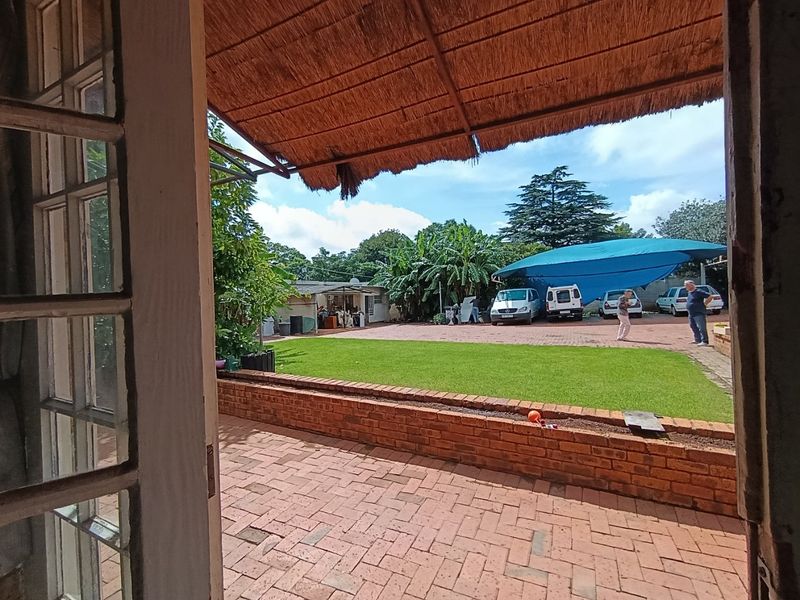 A covered patio area with a thatch roof overlooking a paved backyard, lawn, and garden space.