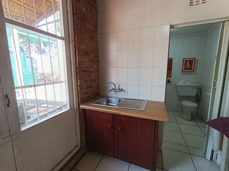 A functional scullery area featuring a stainless steel sink, tiled splashback, and exposed brickwork, with direct access to an adjacent bathroom.