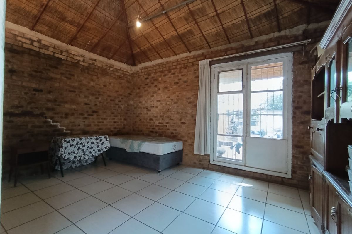 A rustic bedroom featuring exposed brick walls, a high thatched ceiling, tiled flooring, and a glass-paneled door with security bars.