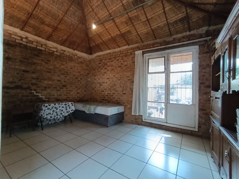 A rustic bedroom featuring exposed brick walls, a high thatched ceiling, tiled flooring, and a glass-paneled door with security bars.