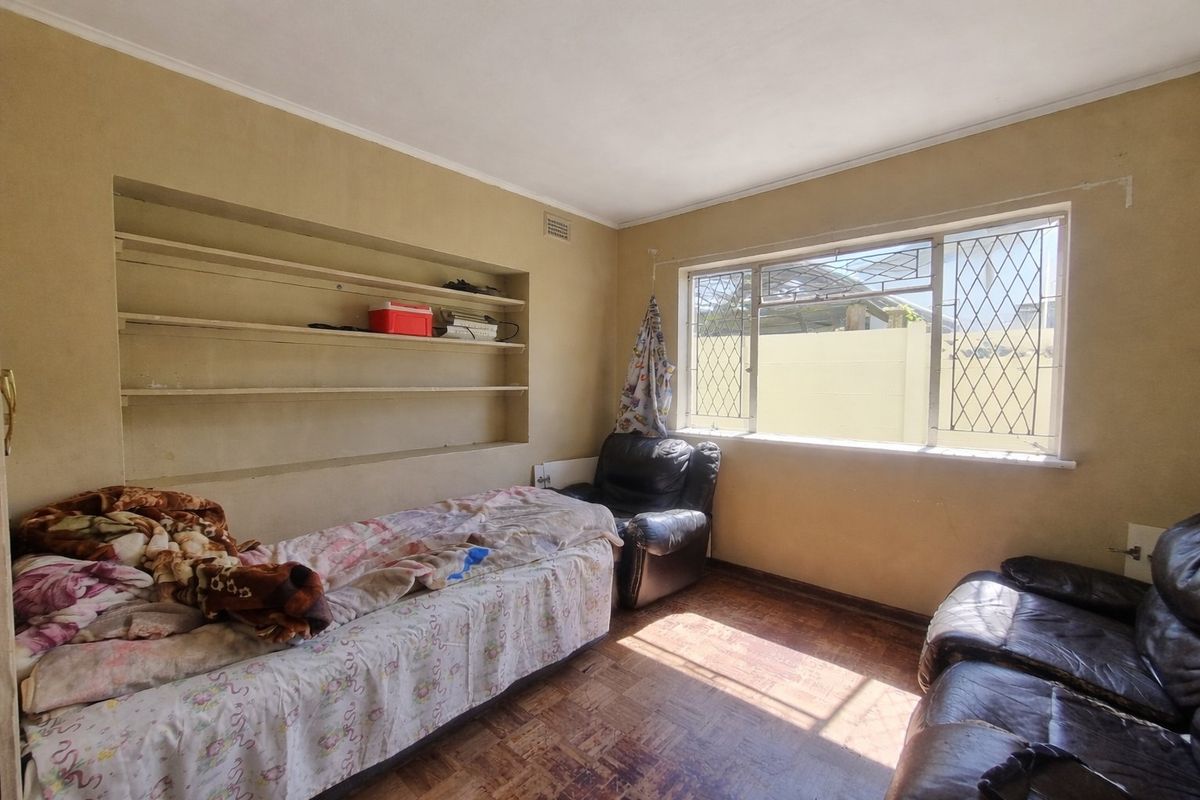 A bedroom featuring parquet flooring, built-in wall shelving, and a window with security bars, offering a functional space with potential for cosmetic updates.