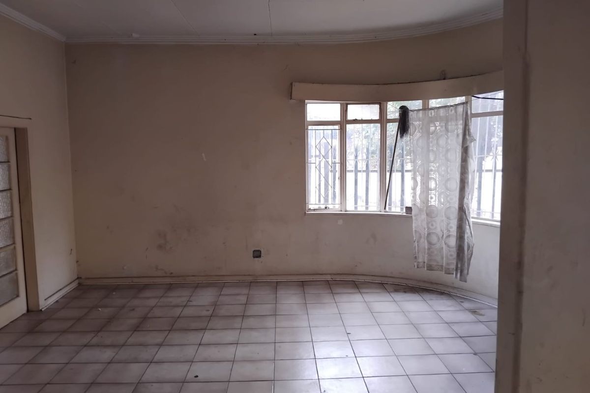 An empty living room featuring a bay window with security bars, tiled flooring, and coving, requiring cosmetic updates to walls and ceiling.