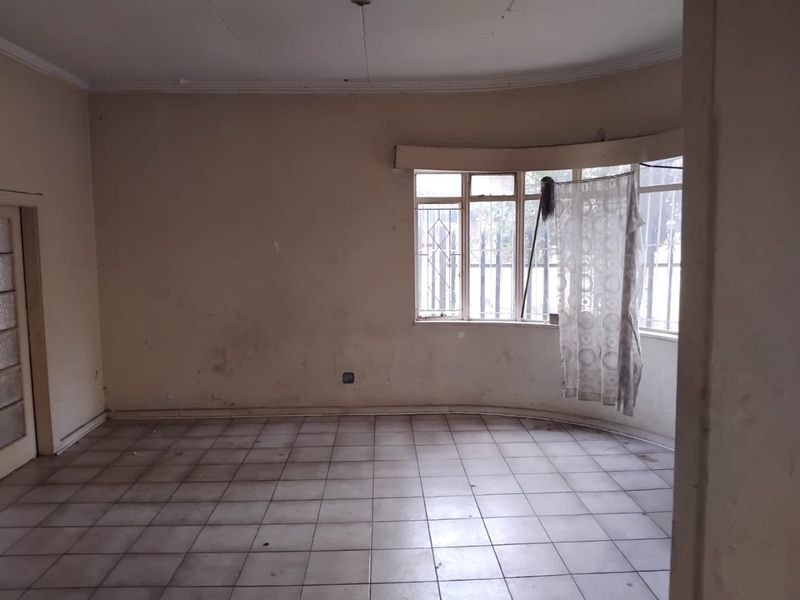 An empty living room featuring a bay window with security bars, tiled flooring, and coving, requiring cosmetic updates to walls and ceiling.