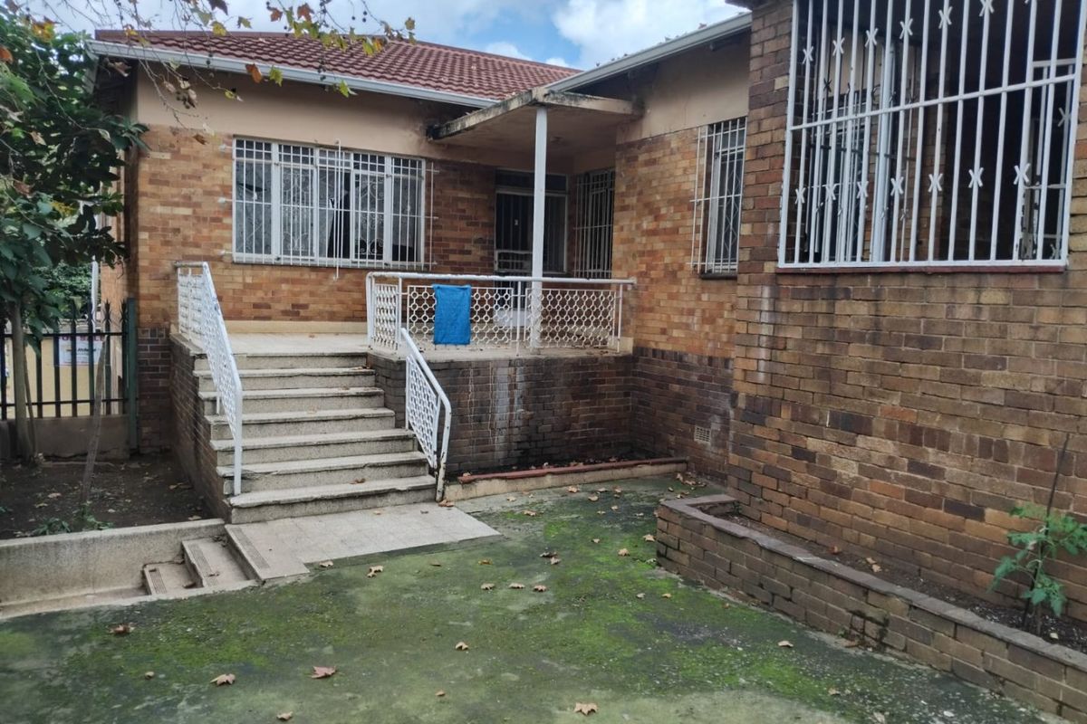 Rear exterior view of a face brick house featuring a raised concrete patio, security bars on windows, and a paved garden area requiring maintenance.