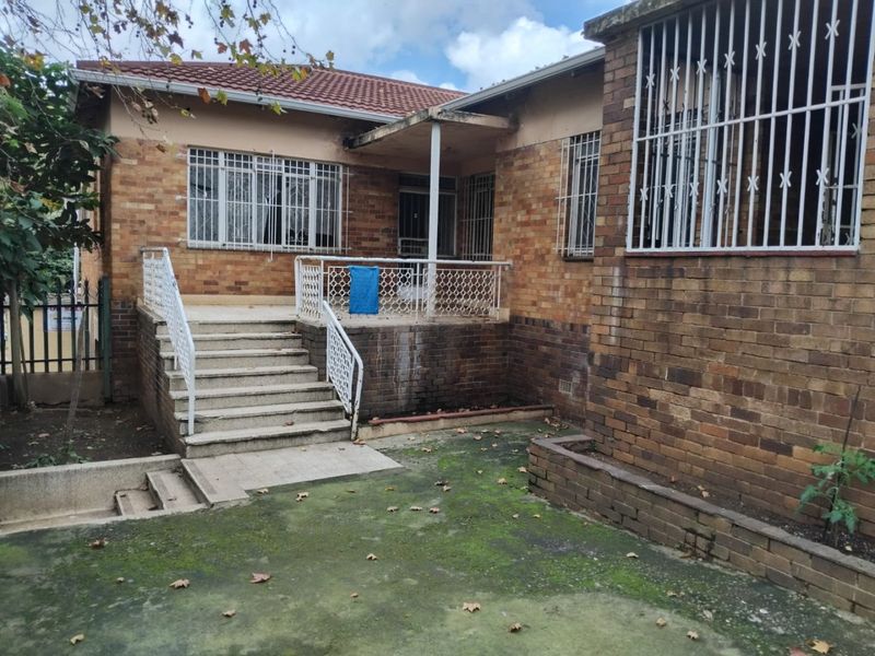 Rear exterior view of a face brick house featuring a raised concrete patio, security bars on windows, and a paved garden area requiring maintenance.