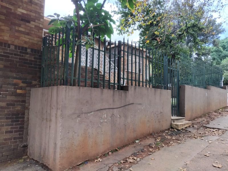 Street-level view of the property featuring a concrete boundary wall, metal security fencing, and a pedestrian gate leading to the brick-faced residence.