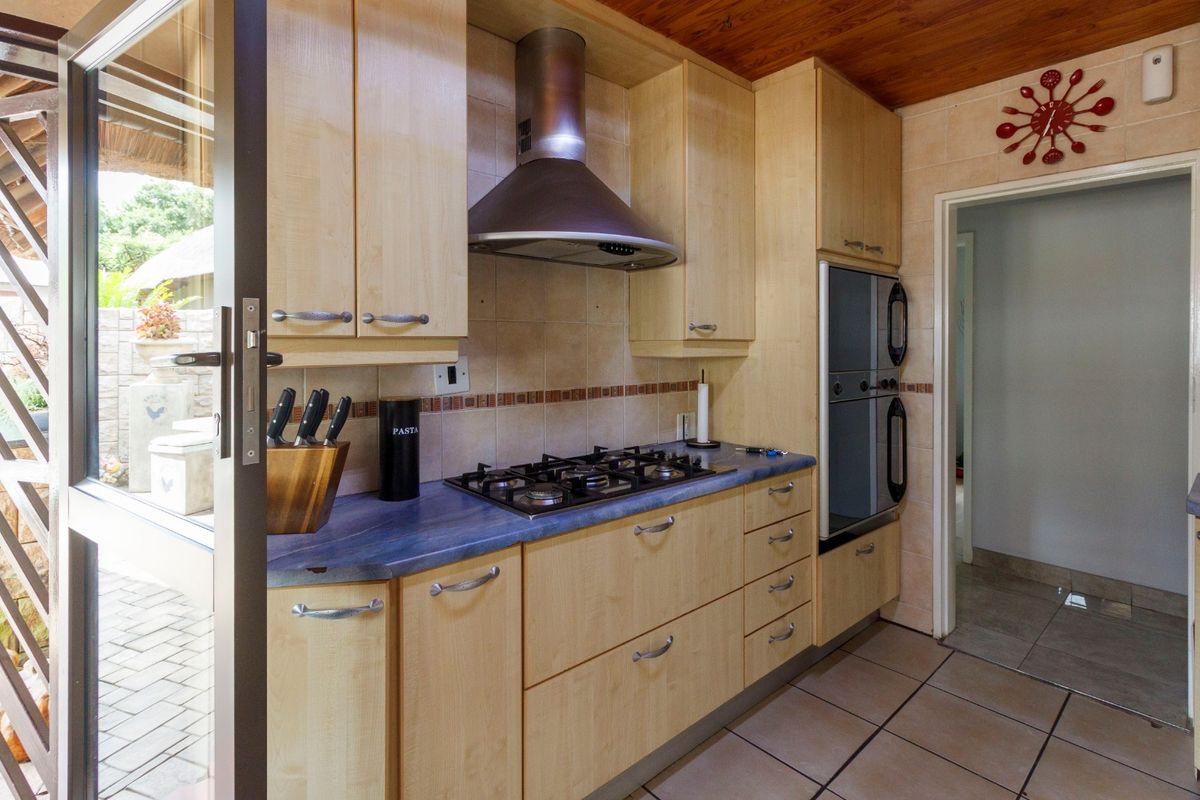 A functional kitchen featuring light wood cabinetry, a gas hob with extractor fan, built-in double oven, and tiled flooring with direct access to the patio.