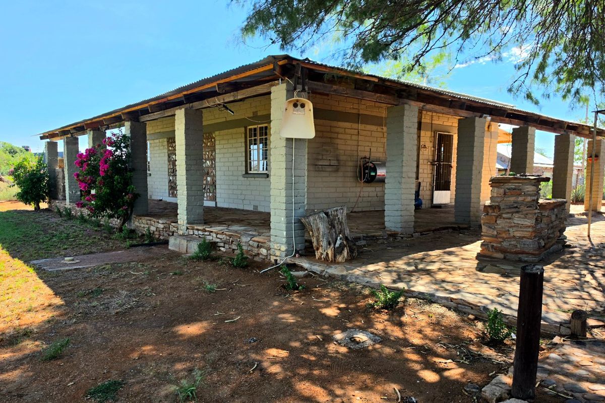 A rustic farm-style dwelling featuring a covered stone patio, exposed brick walls, and a built-in stone braai area under a corrugated metal roof.