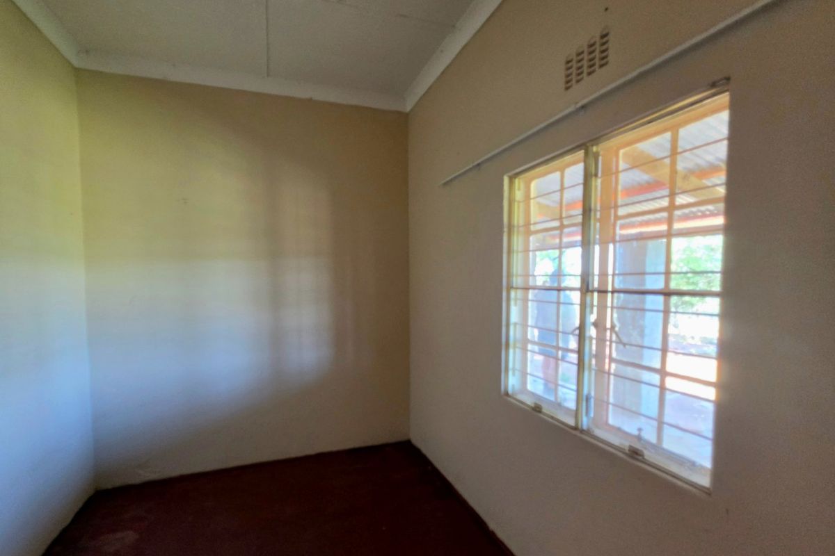 An empty bedroom featuring plain walls, a window with security bars, and a ventilation grille, offering a blank canvas for renovation.