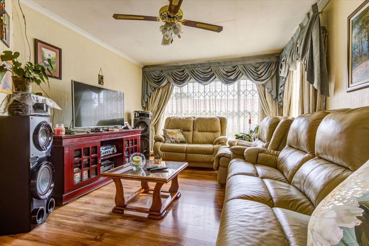 A comfortable living room featuring laminate flooring, a ceiling fan, and large windows fitted with security gates and decorative pelmets.