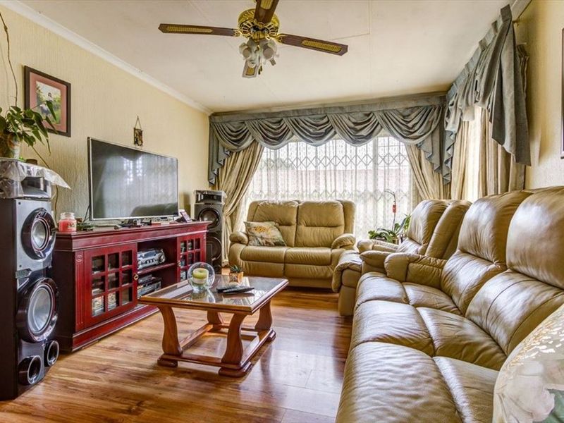 A comfortable living room featuring laminate flooring, a ceiling fan, and large windows fitted with security gates and decorative pelmets.