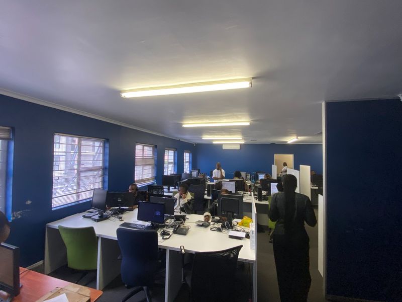 Open-plan commercial office space featuring multiple workstations, fluorescent lighting, and large windows providing natural light.