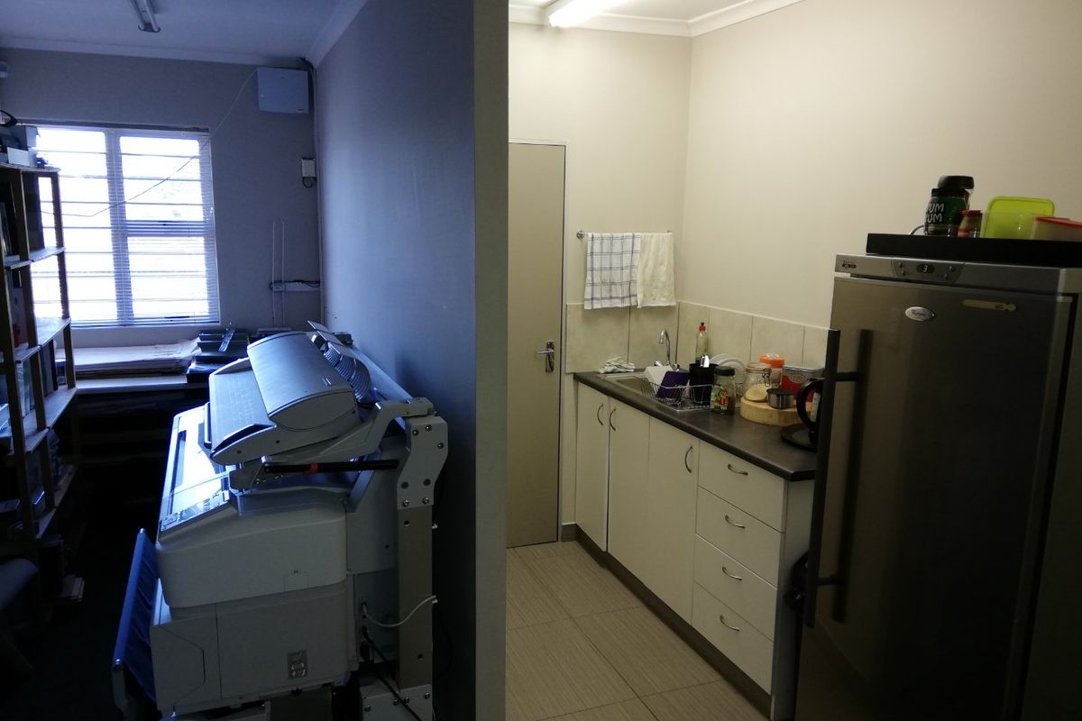 A functional commercial kitchenette area featuring built-in cabinetry, a stainless steel sink, and tiled flooring, adjacent to a workspace.