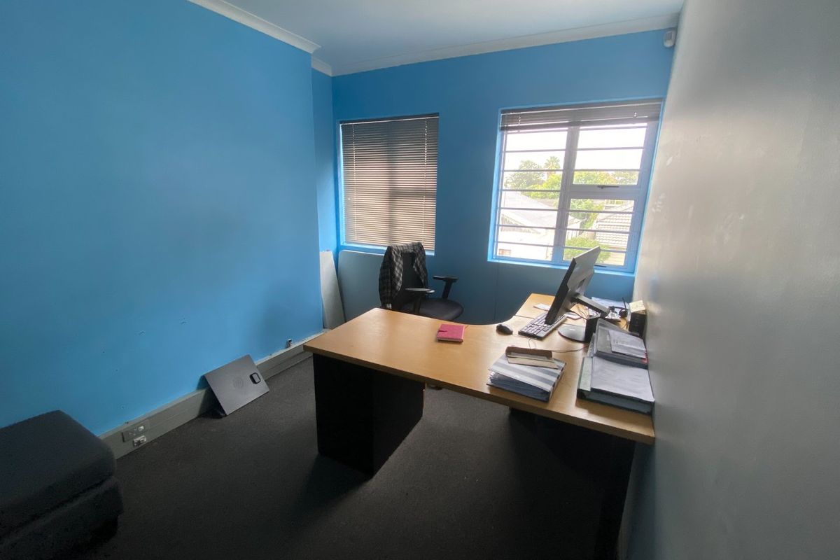 A functional office space featuring blue walls, carpeted flooring, and two windows with fitted blinds, providing natural light for a workspace.