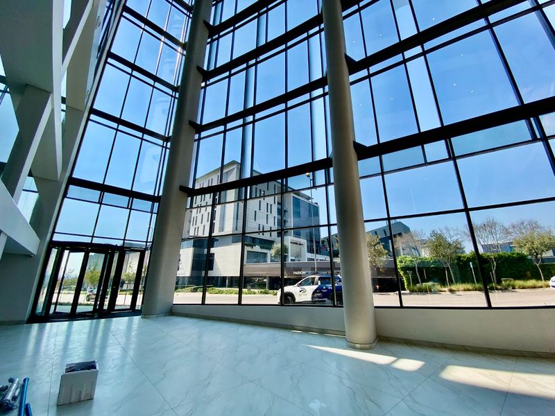 Spacious commercial entrance lobby featuring floor-to-ceiling glass curtain walls, structural columns, and polished marble-look flooring with abundant natural light.