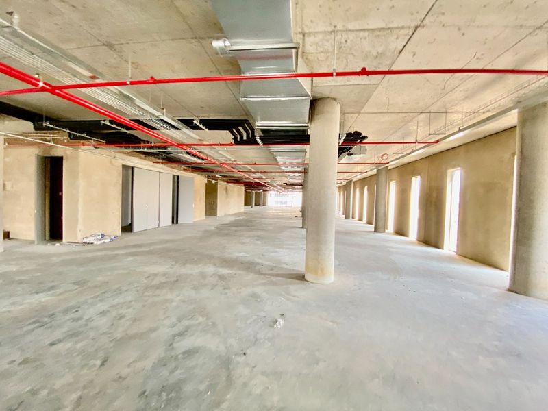 A large, open-plan commercial space in shell condition, featuring exposed concrete ceilings, structural columns, and visible fire suppression and HVAC infrastructure.