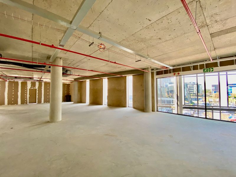 An open-plan commercial office space in shell condition, featuring exposed concrete ceilings, fire sprinkler systems, and large windows with urban views.