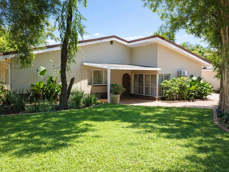 A well-maintained rear exterior view of the house featuring a covered patio, paved pathways, and a lush, established garden with mature trees.