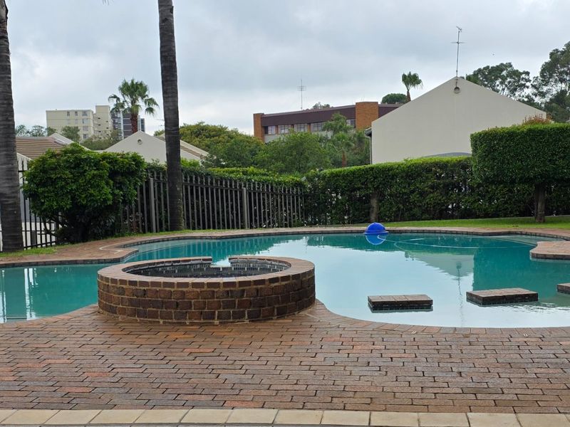 Communal swimming pool area featuring brick paving, a raised brick feature, and established perimeter hedging within a secure townhouse complex.