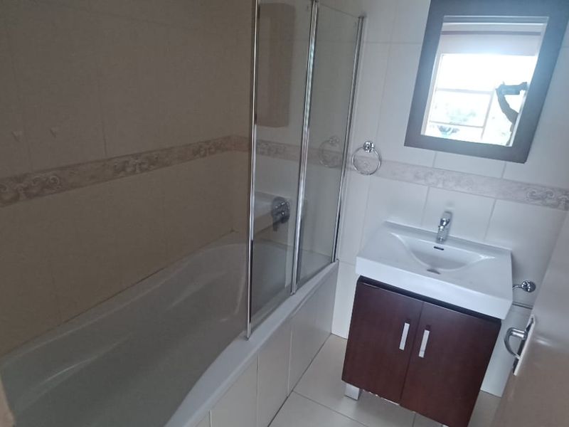 A functional bathroom featuring a bathtub with a glass screen, a vanity cabinet with a basin, and neutral wall and floor tiling.